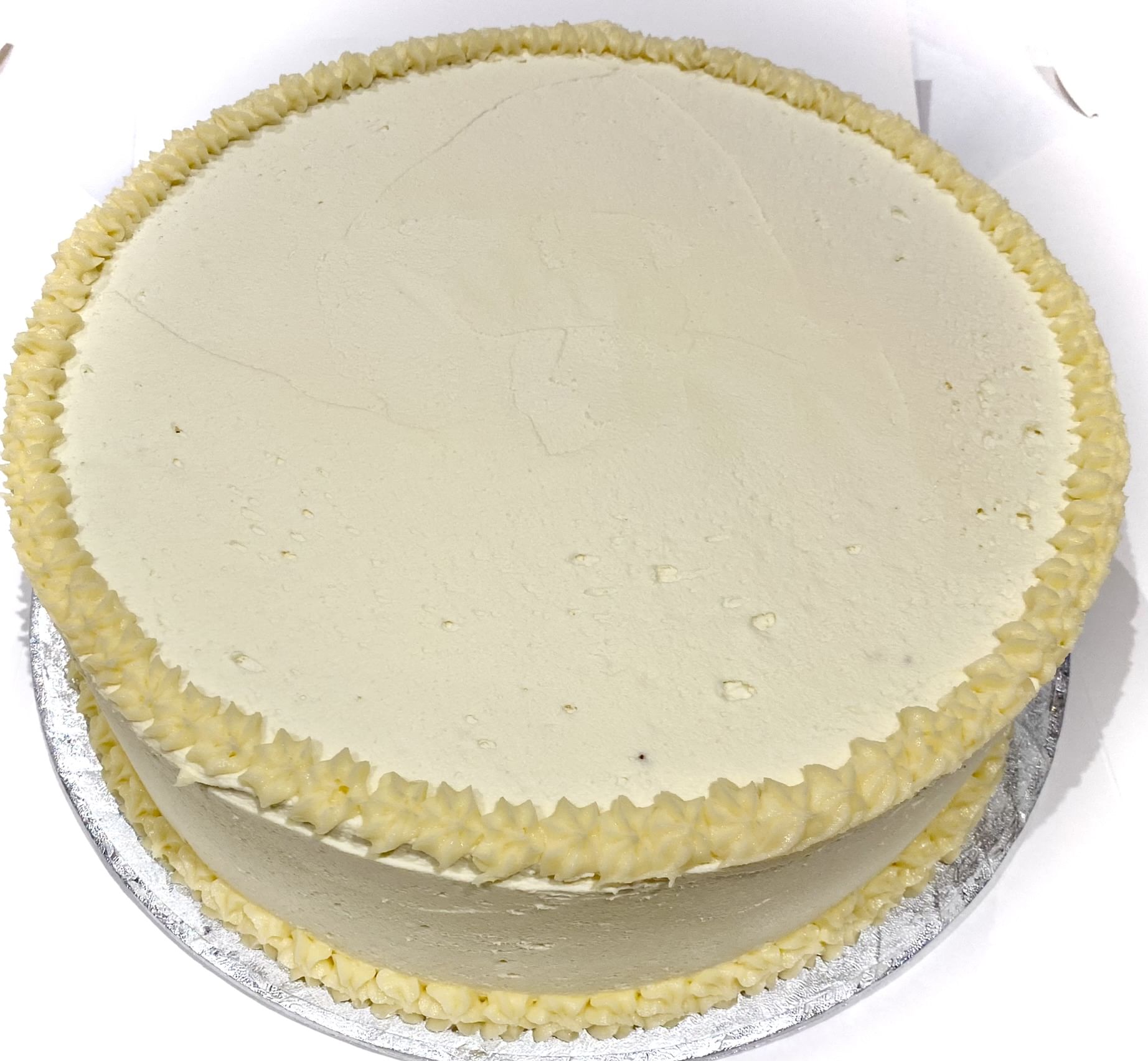 A classic Vanilla Gateaux with smooth white, creamy frosting, decorated with a simple border of piped yellow cream around the top edge. The cake sits elegantly on a silver cake board.